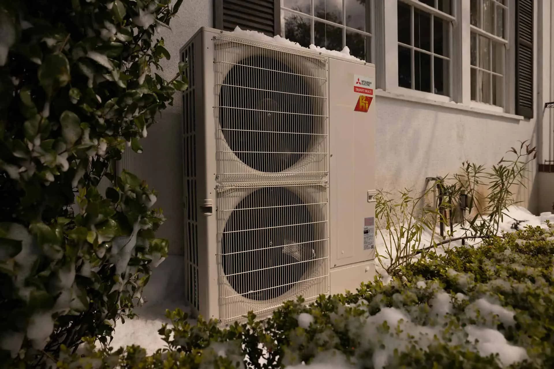 A Mitsubishi heat pump outside a Denver home, the shrubs around it covered in snow.